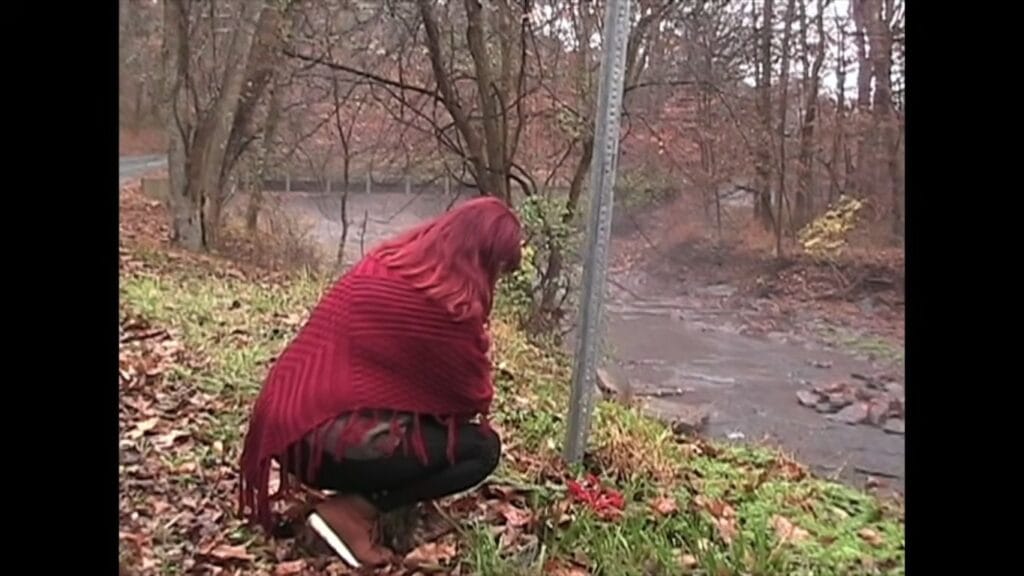 A woman with red hair wearing a knit shawl crouching by a metal signpost near a muddy stream in Tinsman Road.