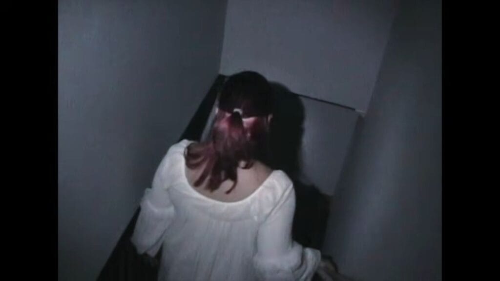 A person in a white nightgown walking down a narrow, shadowy hallway seen from behind in Tinsman Road.