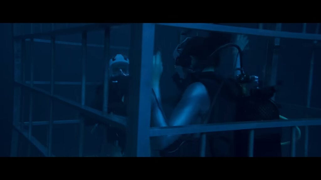 Divers in scuba gear clinging to the metal bars inside a submerged shark cage in Above and Below.