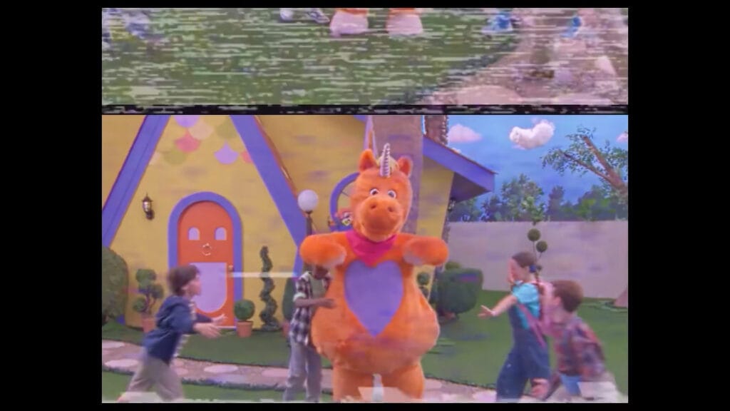 Buddy the orange unicorn mascot standing in front of a colourful playhouse surrounded by children on a retro TV broadcast in Buddy.