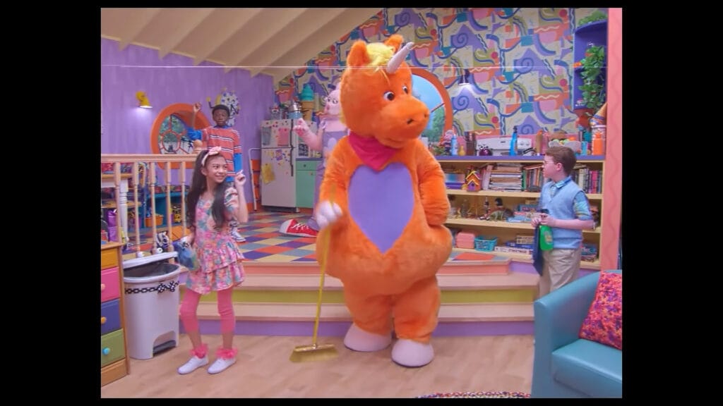 Buddy the orange unicorn sweeping the floor with a broom alongside smiling child cast members on a brightly coloured studio set in Buddy.