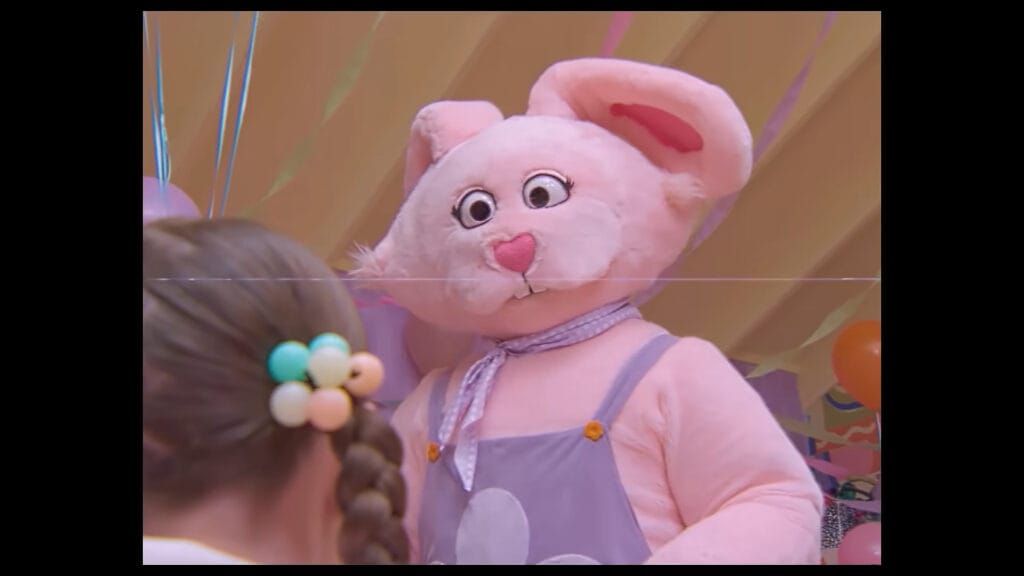 A pink costumed rabbit mascot with wide uncanny eyes staring down at a young girl in Buddy.