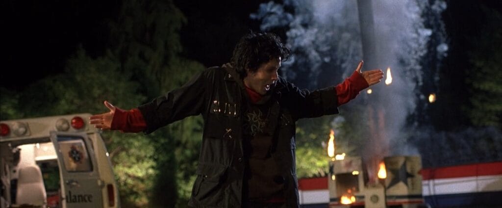 Ian standing outside at night with outstretched arms shouting near an ambulance and rising flames in Final Destination 3.