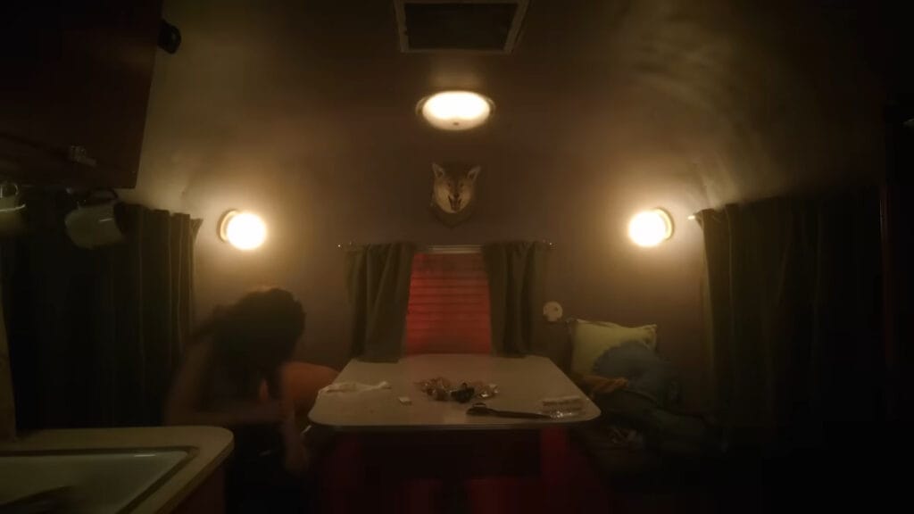 Interior wide shot of a dimly lit vintage Airstream camper trailer with glowing wall sconces and a mounted coyote head in Hellcat.