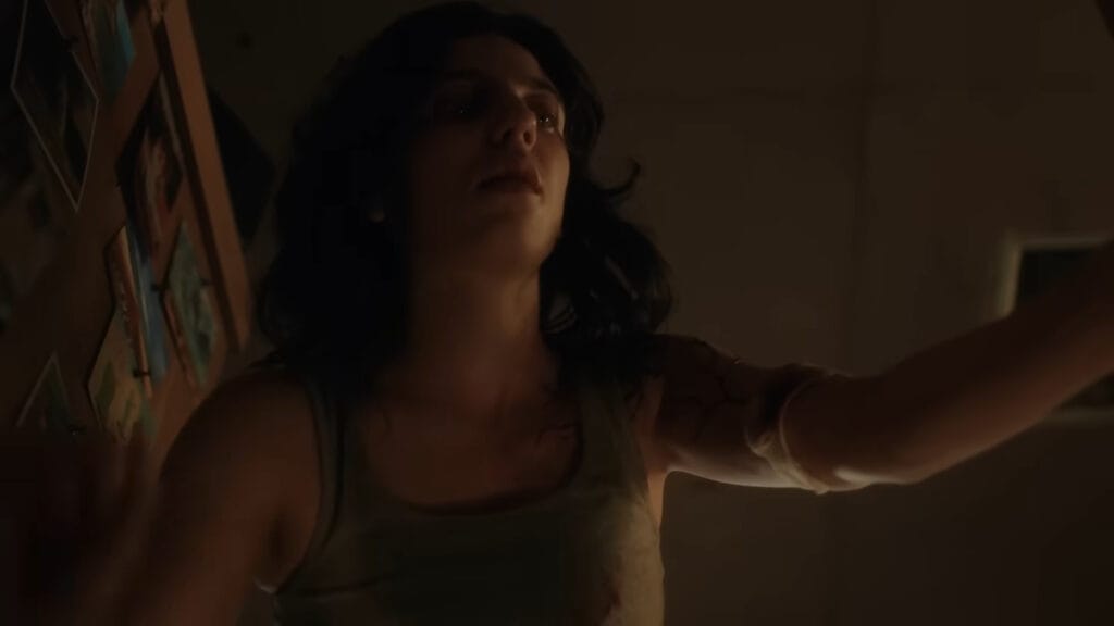 Dakota Gorman as Lena with dark veins on her shoulder looking upwards with mouth open inside a dim trailer in Hellcat.