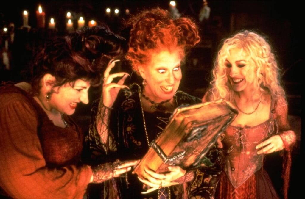 The Sanderson sisters—Mary, Winifred, and Sarah—smiling down as they open their glowing spellbook in Hocus Pocus (1993).