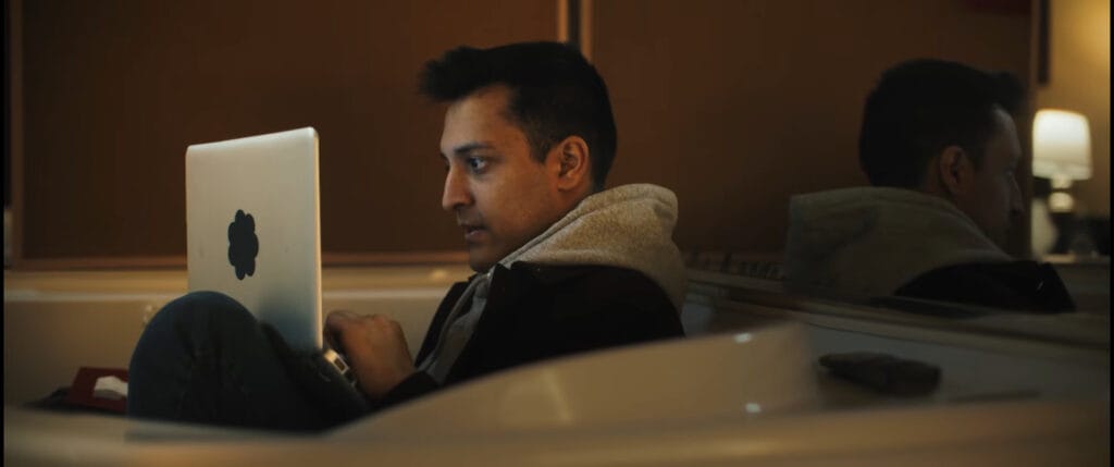 Rushabh Patel as Rian sitting in an empty bathtub working intently on a laptop inside a motel bathroom in I Know Exactly How You Die.