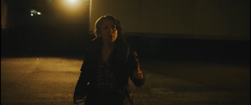 Woman walking through a dim, street-lit outdoor lot at night looking uneasy in I Know Exactly How You Die.