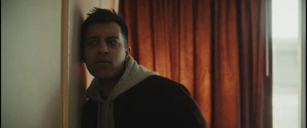 Rushabh Patel as Rian leaning his head against a wall listening through to the adjoining room with a frustrated expression in I Know Exactly How You Die.