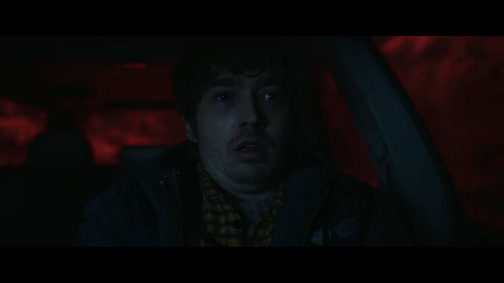 Austin Abrams as Bryan staring through a car windshield with a shocked, panicked expression bathed in red brake lights in Resident Evil.