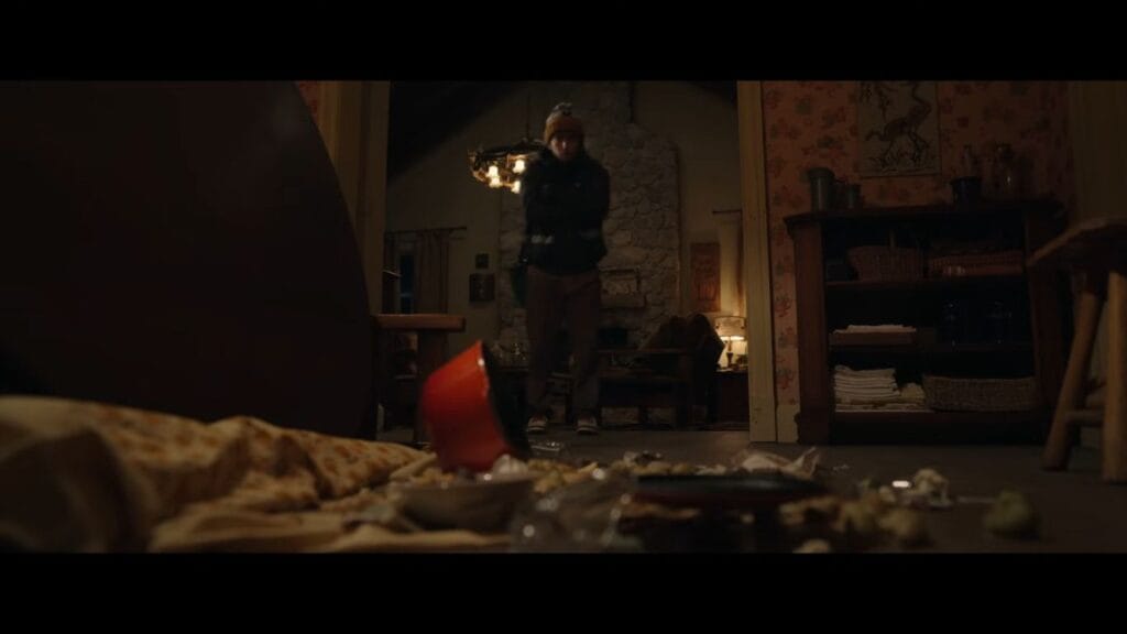 Austin Abrams as Bryan standing cautiously in the doorway of a messy, ransacked living room in Resident Evil.