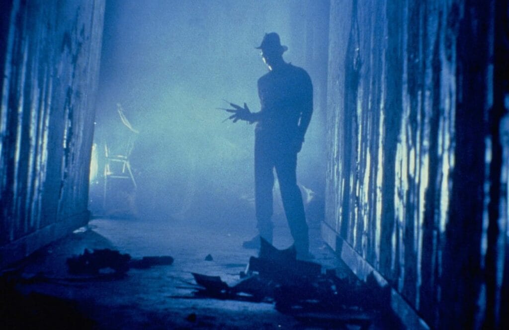 Silhouette of Freddy Krueger with his razor-glove raised standing down a dark, misty corridor lit with cold blue light in A Nightmare on Elm Street 3: Dream Warriors.