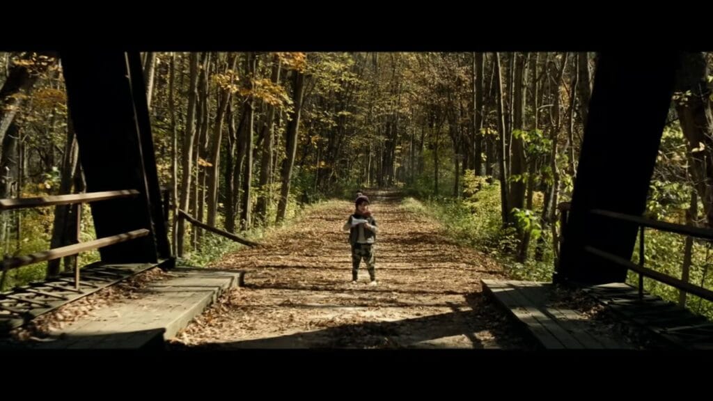 A small boy standing alone on a leaf-covered railway bridge holding a toy spaceship in A Quiet Place.