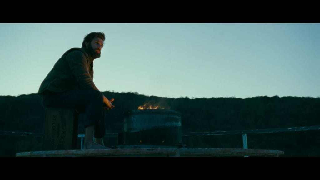 John Krasinski as Lee sitting barefoot beside a fire brazier on top of a farm silo at dusk in A Quiet Place.