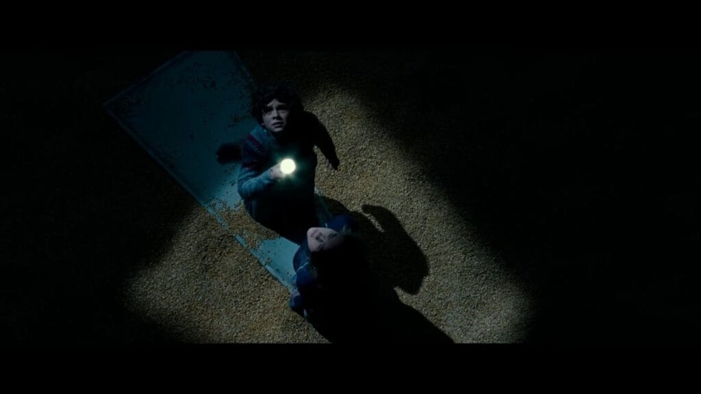 Overhead shot of two children trapped atop a metal sheet on sinking grain inside a dark silo in A Quiet Place.