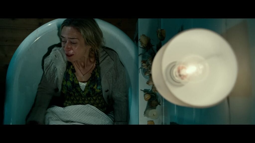 Emily Blunt as Evelyn weeping silently while hiding in a bathtub beneath a hanging lamp in A Quiet Place.