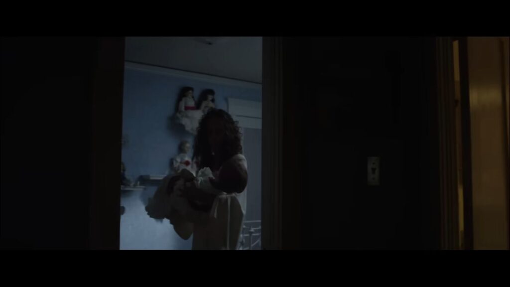 A woman in a white nightgown holding an infant in a dimly lit doorway with dolls visible on shelves in Annabelle.