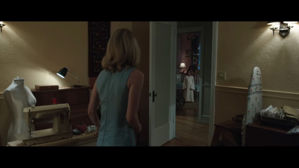 Mia standing in a sewing room looking through a doorway toward a shadowy figure in a white gown in Annabelle.
