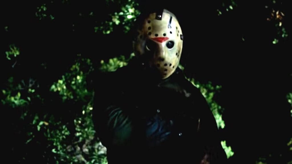 Jason Voorhees standing motionless wearing his iconic hockey mask in the dark woods in Friday the 13th Part VI: Jason Lives.