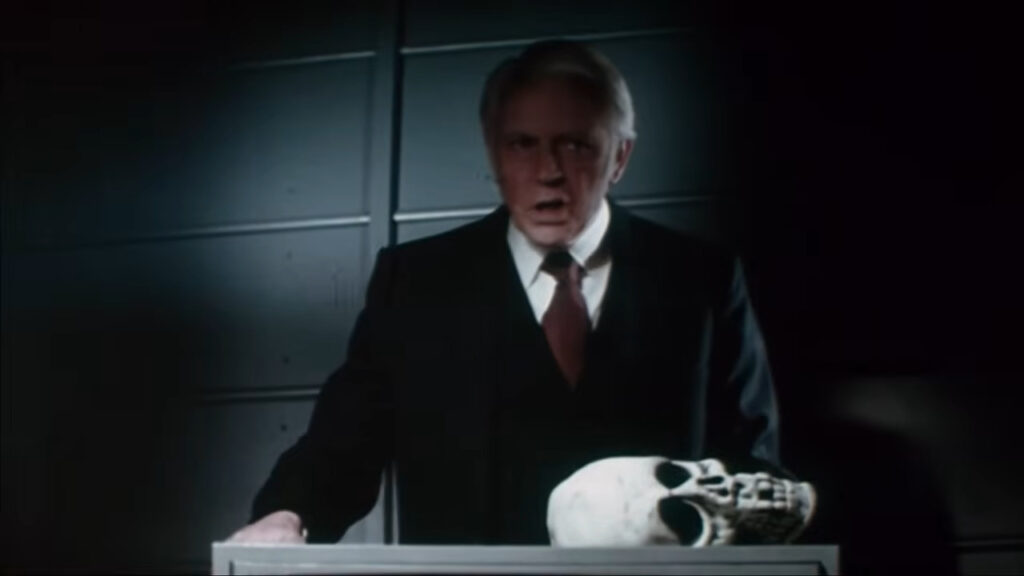 Dan O'Herlihy as Conal Cochran standing menacingly in a dark suit behind a podium with a human skull in Halloween III: Season of the Witch.