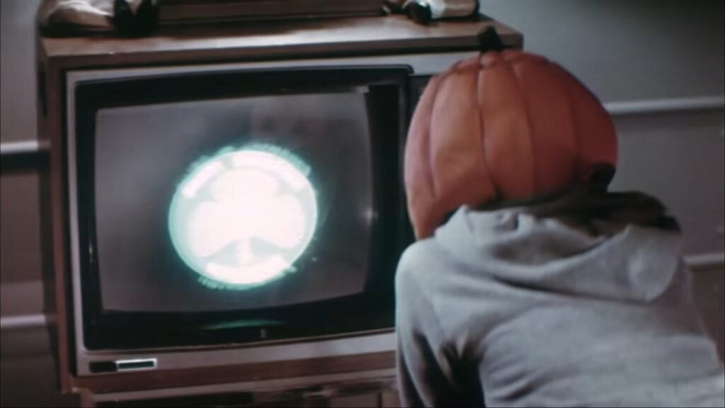 A child wearing a Silver Shamrock jack-o'-lantern mask watching a glowing commercial on a CRT television screen in Halloween III: Season of the Witch.