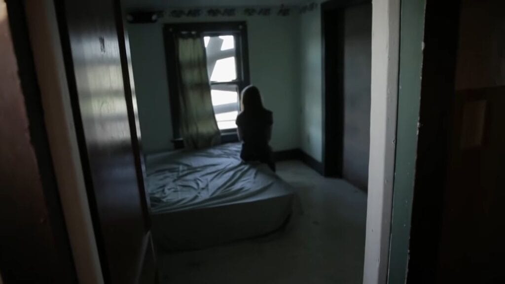 A person sitting alone on a bare mattress inside a dimly lit, rundown room in Hell House LLC.
