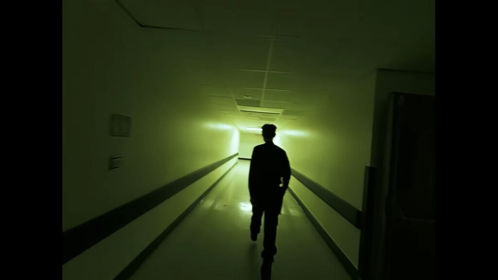 Silhouette of a security guard walking down a long, dimly lit green hospital corridor in Infirmary.