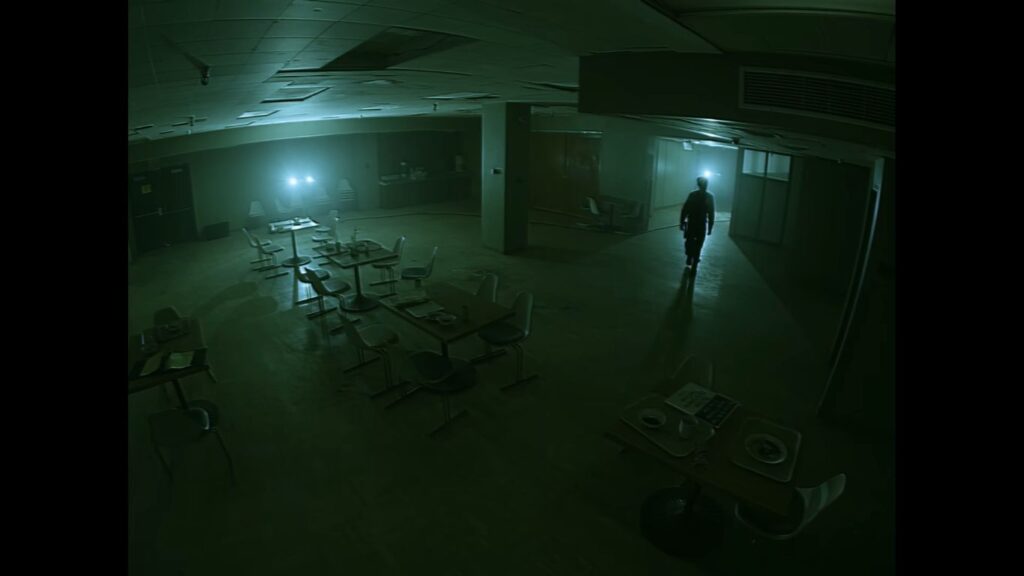 High-angle CCTV security camera view of a dark, abandoned hospital cafeteria with a lone figure walking in the distance in Infirmary.