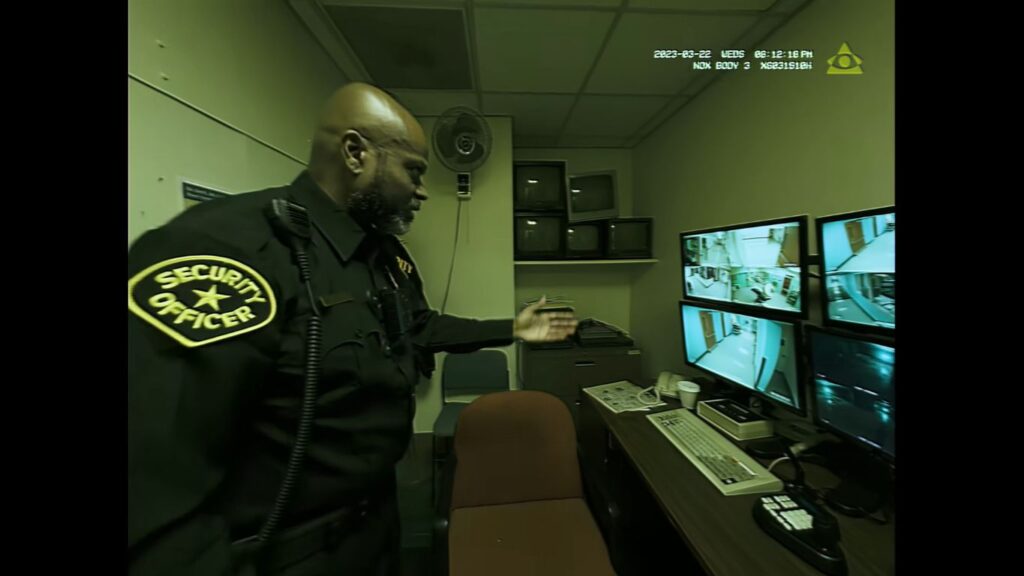 A senior security guard gesturing toward multiple surveillance monitors in a security office in Infirmary.