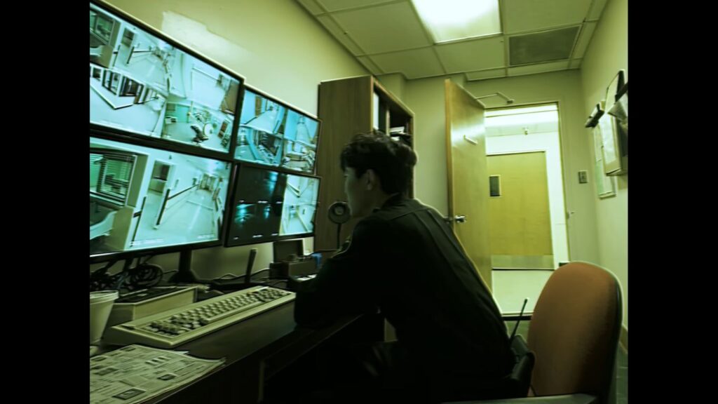 Paul Syre Lee as Edward seated at a desk monitoring multiple security screens in Infirmary.