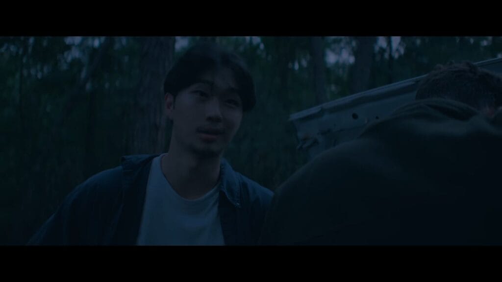 Phinehas Yoon looking weary and concerned while talking beside a vehicle in the dark woods in It Ends.