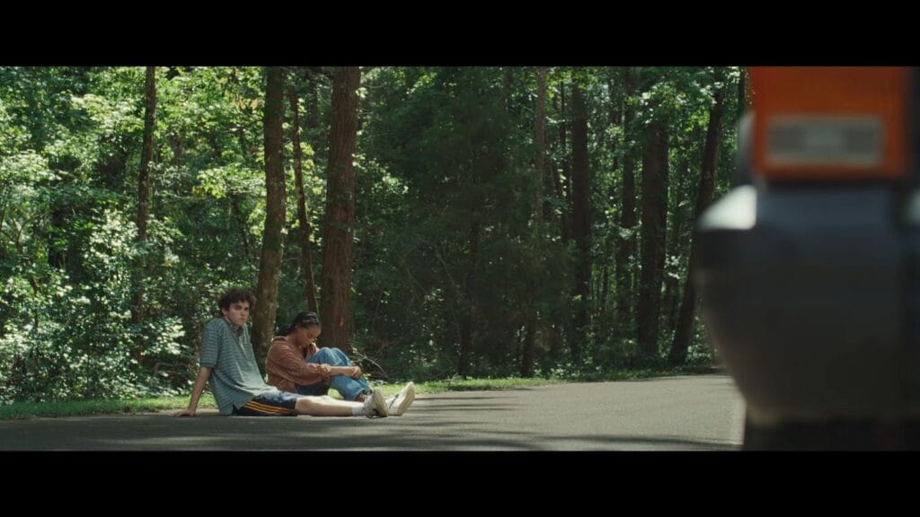 Two exhausted young adults sitting on an empty asphalt road beside a dense forest in It Ends.