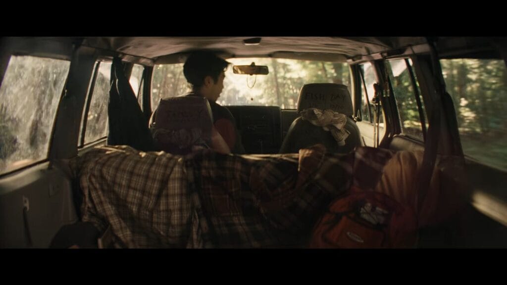 Interior view from the back of an SUV showing luggage and the driver looking out at a forest road ahead in It Ends.