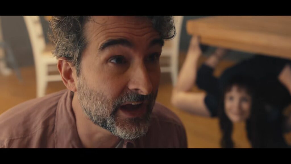 A man looking panicked in the foreground while Jessica Chastain's character creeps upside down underneath a table in Other Mommy.