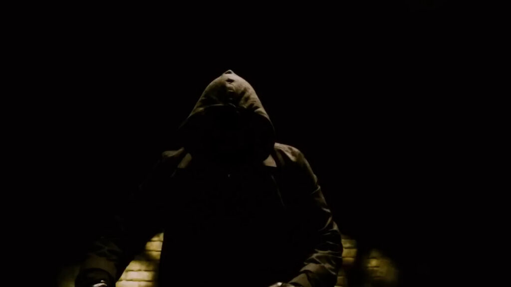A hooded figure sitting in the dark in Saw IV.
