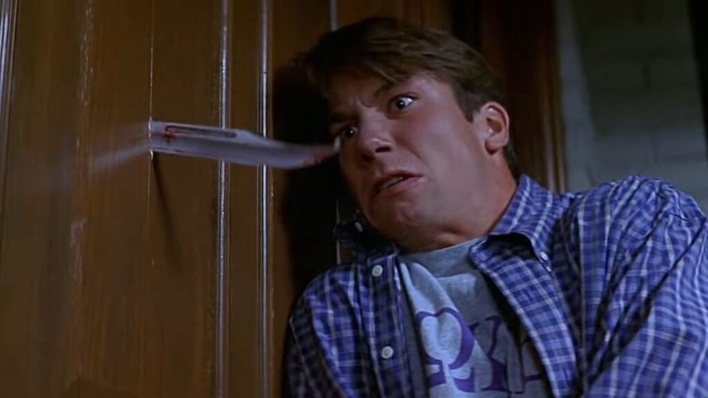 Jerry O'Connell as Derek reacting in terror as a bloody knife blade punches through a wooden door in Scream 2.