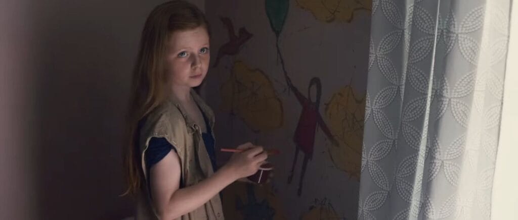A young girl with long reddish hair holding a crayon and painting directly onto a bedroom wall in Sinister.