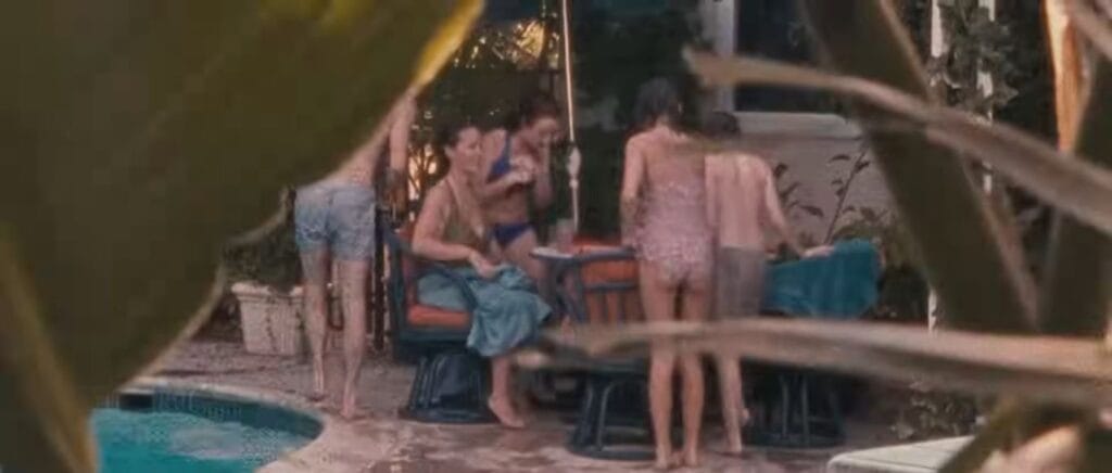 Grainy Super 8 film footage viewed through foliage showing a family relaxing by a backyard swimming pool in Sinister.