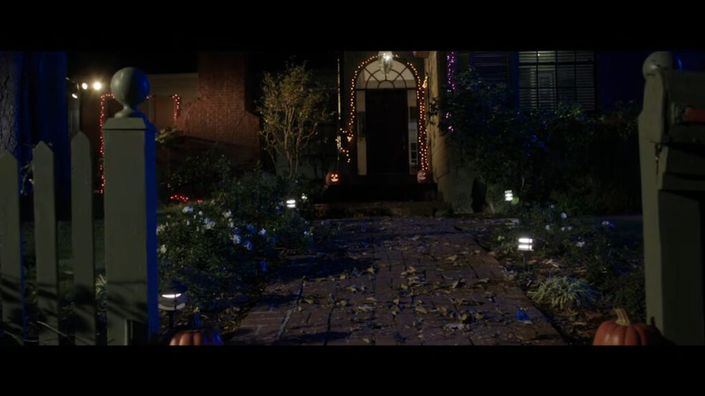 A brick walkway lined with glowing jack-o'-lanterns and lights leading to a decorated suburban house at night in Tales of Halloween.