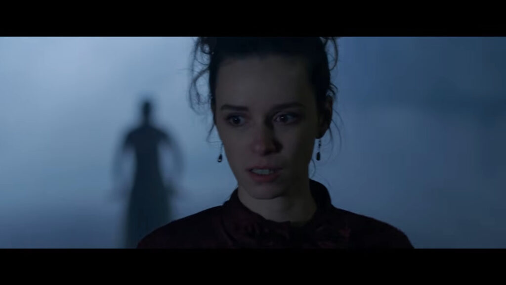 Alex Essoe looking terrified as an ominous shadowy figure looms in the misty background in the Grim Grinning Ghost segment of Tales of Halloween.