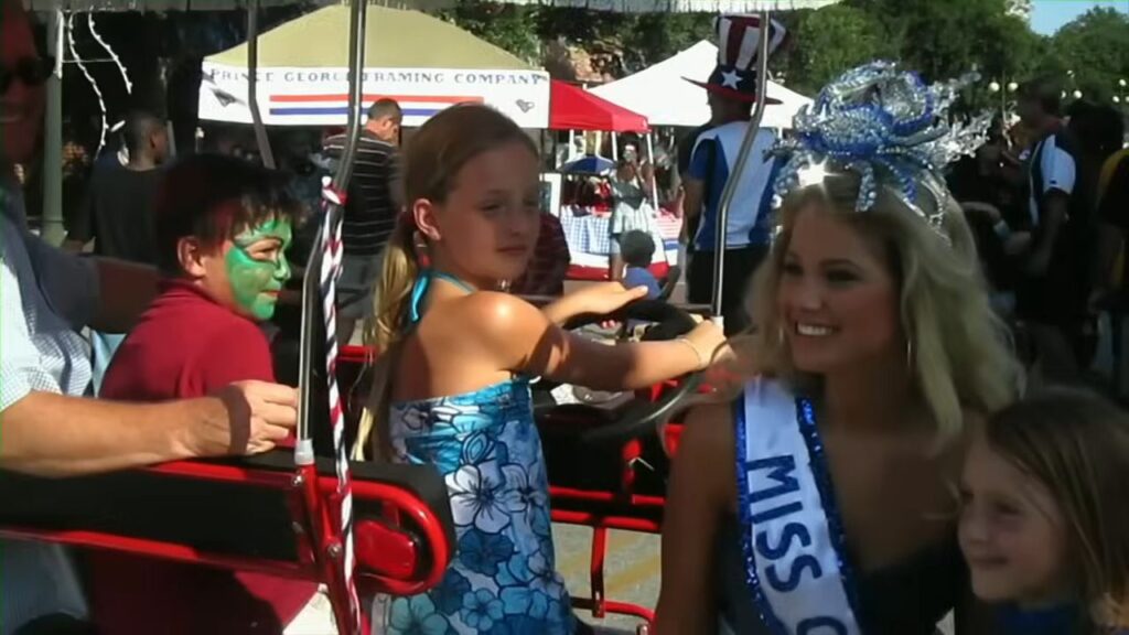 A beauty pageant winner wearing a sash and crab tiara seated next to young girls at a parade in The Bay.