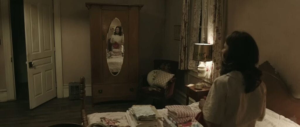 Carolyn folding laundry on a bed while looking at an oval mirror on a wardrobe door in The Conjuring.