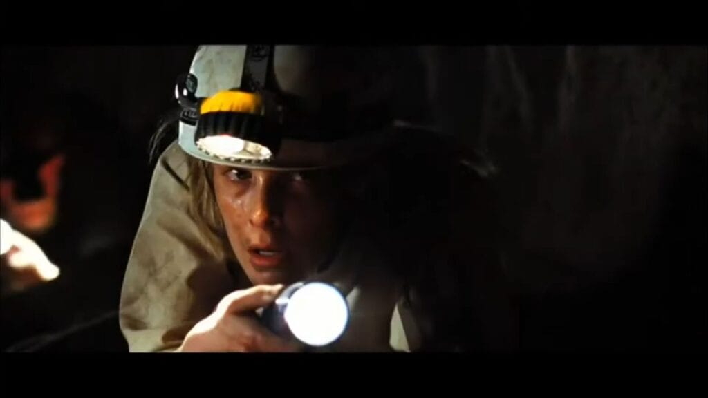 A caver wearing a miner helmet crawling through a dark cave while aiming a flashlight forward in The Descent: Part 2.