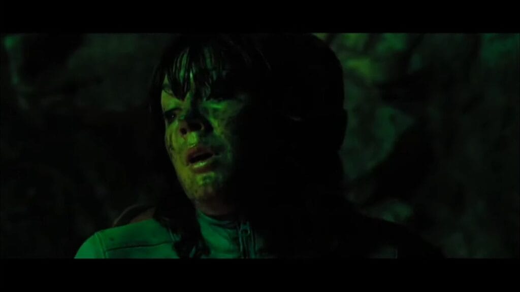 A woman covered in grime and mud looking anxious under eerie green lighting inside a cavern in The Descent: Part 2.