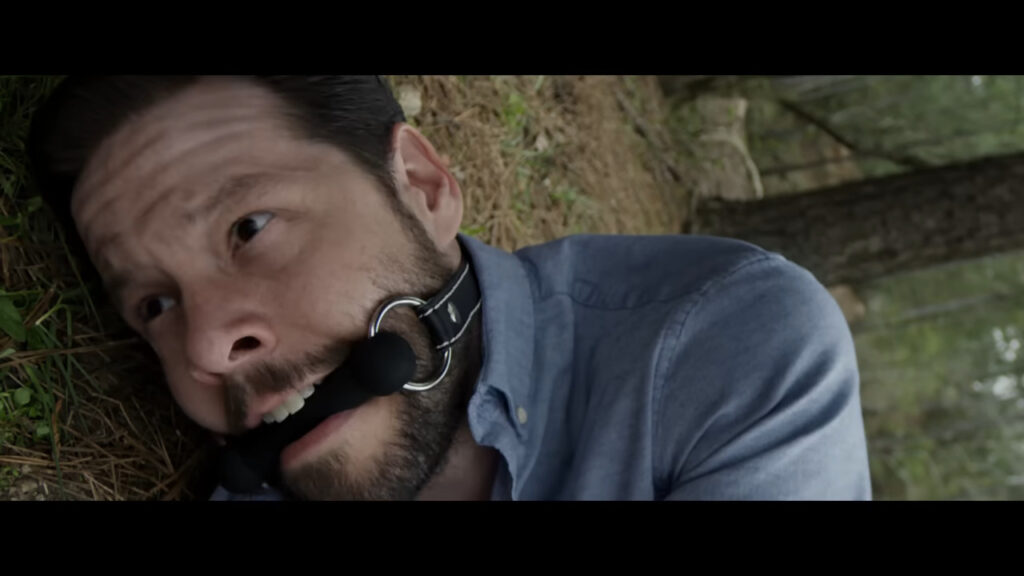 Ike Barinholtz as Staten Island lying on the forest ground gagged with a large black ball gag in The Hunt.