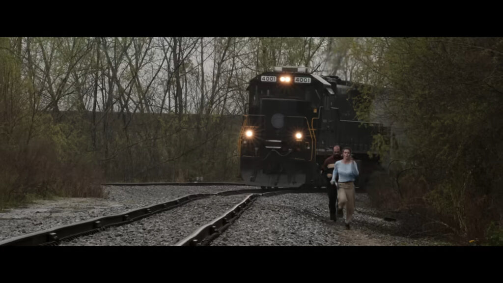 Crystal and Gary desperately running alongside train tracks as a black freight locomotive approaches behind them in The Hunt.