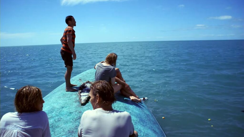 A stranded group sitting and standing atop the overturned hull of their capsized boat in open water in The Reef.