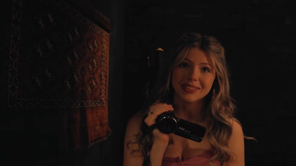 A young woman smiling while holding a digital camcorder in a dimly lit room in V/H/S/Mixtape.