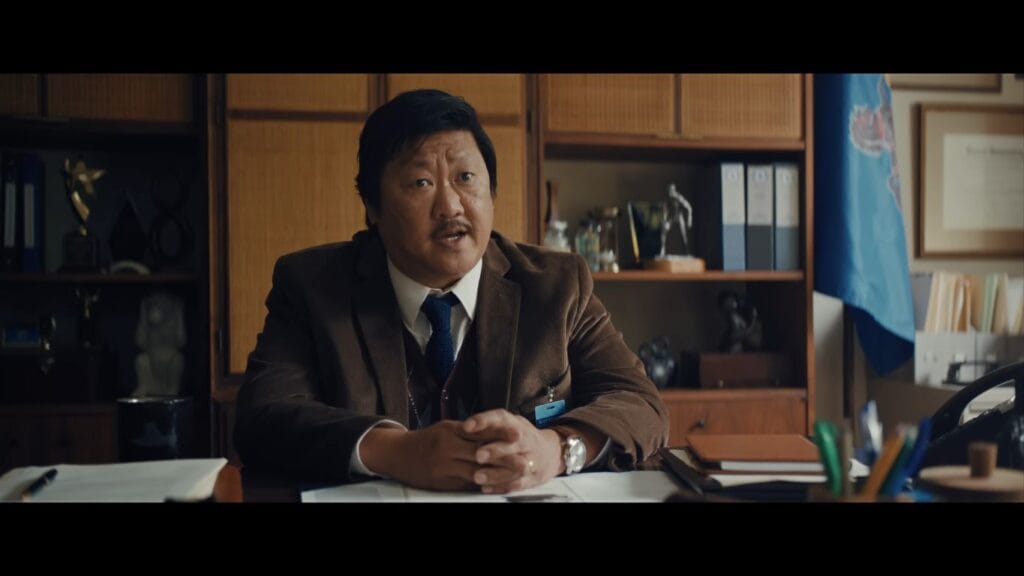 A stern school official in a brown corduroy blazer sitting behind an office desk in Weapons.