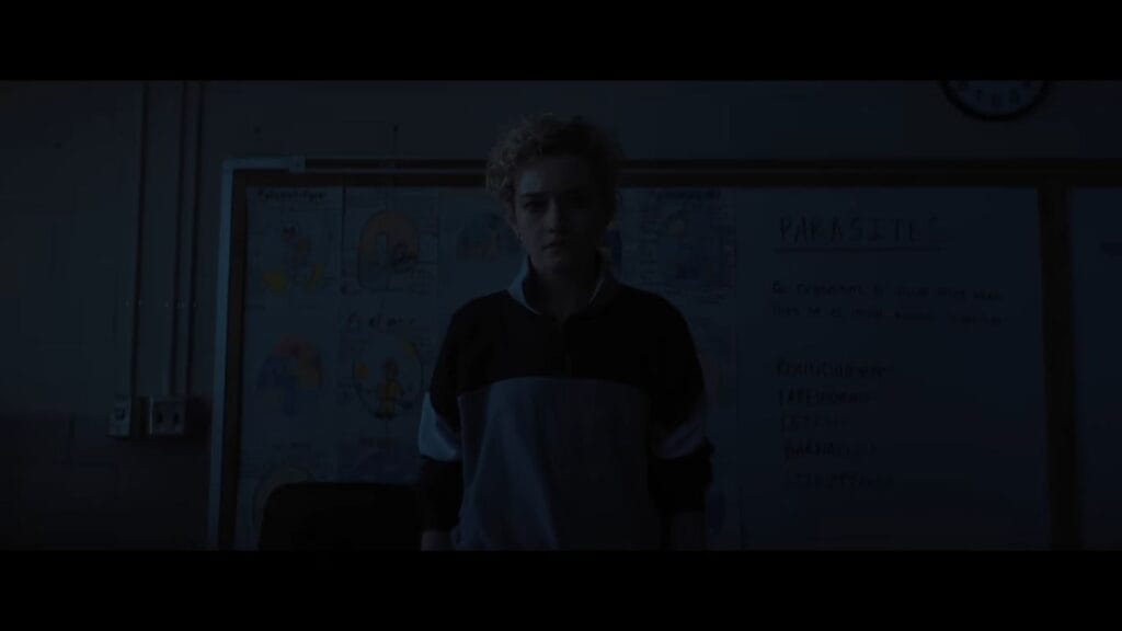 Justine standing alone in a dimly lit classroom in front of a chalkboard detailing parasites in Weapons.
